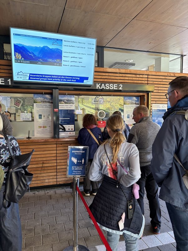 Turisti čekaju kupnju karata za autobuse koji voze prema vrhu, a digitalni ekran prikazuje sliku uživo s planine Kehlstein